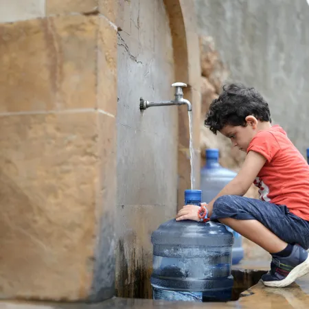 a boy squats in front of a water tap which is filling up a gallon bottle with water.