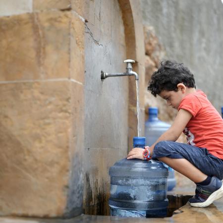 a boy squats in front of a water tap which is filling up a gallon bottle with water. 