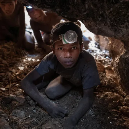 A young person wearing a headlamp crawls through a narrow, rocky cave. Behind them, others follow. The atmosphere is tense and focused.