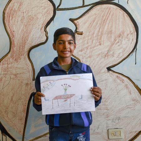 A boy holds a drawing up. It is made on paper and with colour pencils, showing a table with a vase and flowers on it. The boy is looking at the camera smiling. 