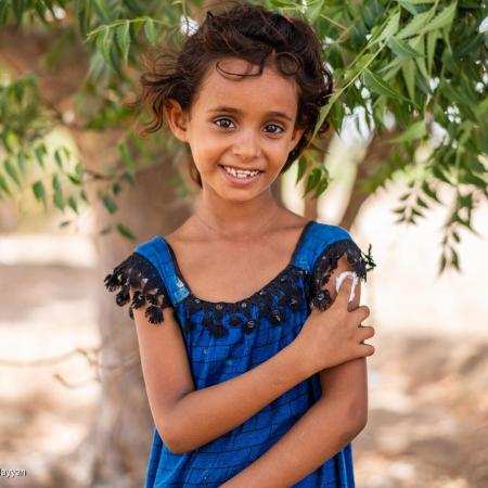 A girl in a blue dress stands under a tree smiling while holding onto a her arm where she has just been given a vaccine shot. This photo was taken in Yemen in 2022. 