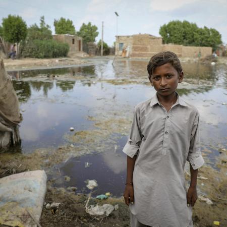A child looks into the camera, behind him are a flooded field and half submerged homes. 