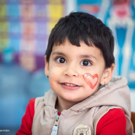 A child with a red heart painted on their face looks at the camera and smiles. They are sitting in a classroom setting. This photo was taken in Bosnia and Herzegovina in 2022.. 