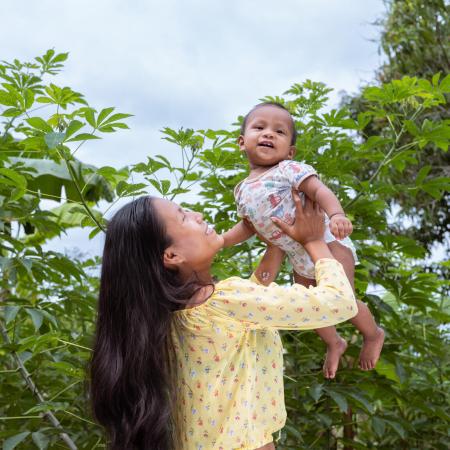 A woman hold up a baby in her arms above her head, behind her are many trees.