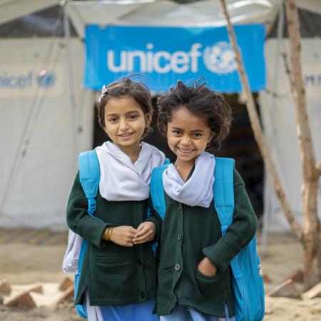 Two young girls wearing blue UNICEF backpacks smile at the camera while standing in front of a UNICEF-funded Temporary Learning Centre.  