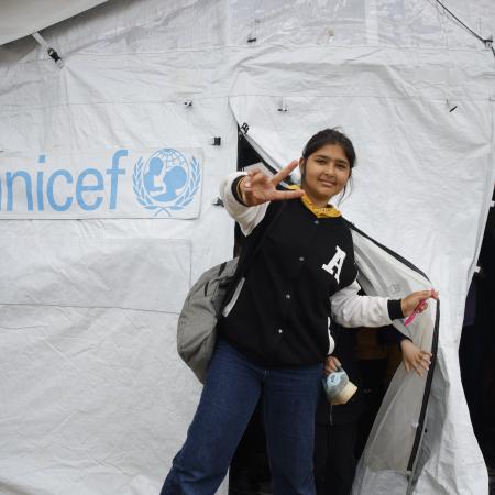 A girl exits a UNICEF tent making the peace gesture with her hand and a bag pack slung over her shoulder.