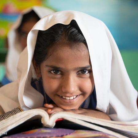 In a classroom in Pakistan, a young girl sitting at her desk smiles with an open book in front of her.  