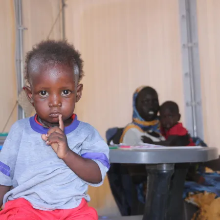 A small child with a serious expression puts a finger to their lips, sitting indoors. In the background, a woman holds another child at a table.
