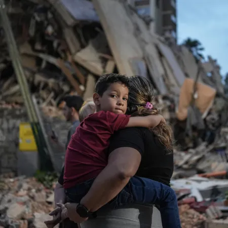 Un enfant est porté par un adulte devant un immeuble effondré.