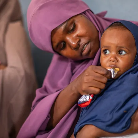 A caring mother in a purple headscarf feeds a young child in a navy hijab, who eats from a snack packet in her arms.