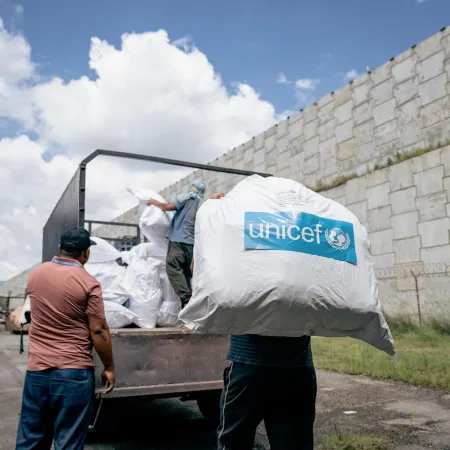 Workers load a large sack with the UNICEF long on it, nto a truck beside a stone wall.