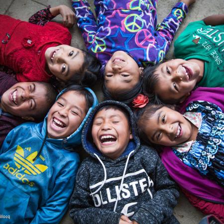 Six children lying on the floor in a circle with their heads touching in. They are looking up at the camera and laughing. 