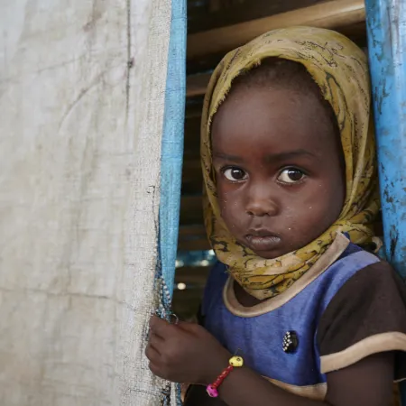 A young child with a serious expression, wearing a yellow headscarf and blue shirt, peeks from behind a curtain