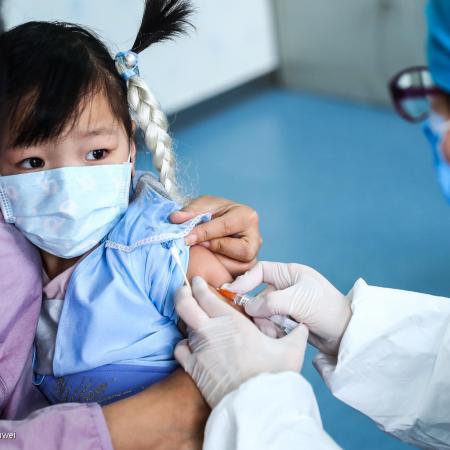 A girl sits in her mother's lap while a health worker administers a vaccine. Both the child and the doctor are wearing masks, the mother's face is not visible. It is a clinic setting. This photo was taken in China in 2020. 