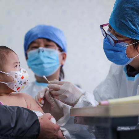 Baby wearing mask looking a doctor in a cap and mask holding a cotton swab with gloved hands