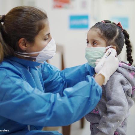 A health worker wearing PPE checks the temperature of a young girl with a digital thermometer. Both are wearing masks. This photo was take in Lebanon in 2020.