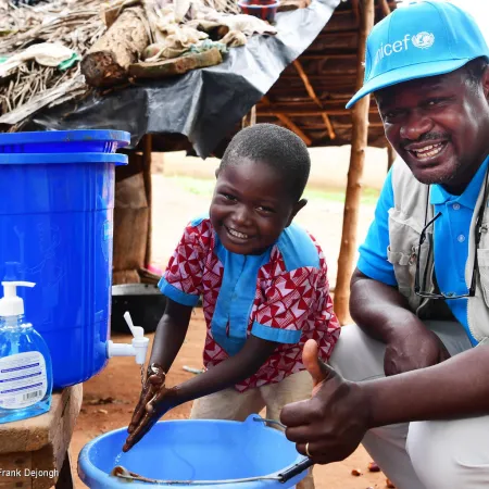 A man and a child squat in front of a bucket with a tap, the boy is washing his hands and the man is giving a thumbs up sign. The man is wearing a UNICEF cap and vest. 