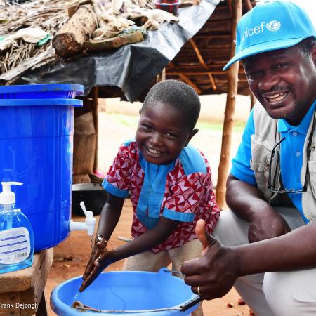 A man and a child squat in front of a bucket with a tap, the boy is washing his hands and the man is giving a thumbs up sign. The man is wearing a UNICEF cap and vest.