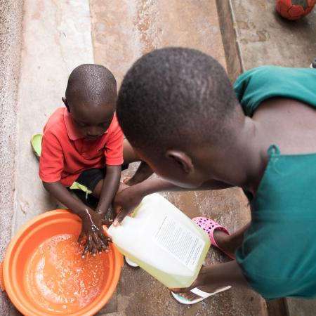 Sharon helps Jonathan to wash his hands with the water and soap mixture at the entrance of their house. In Uganda, the COVID-19 pandemic and lock down has left many families with no option but to stay home and keep stay safe. 