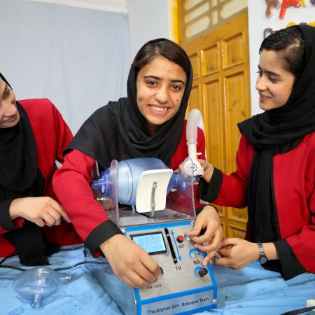 Three girls in red uniforms and black headscarves stand around a desk with a ventilator on it. 