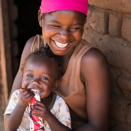 A smiling woman embraces a giggling child holding a packet of peanut paste. 