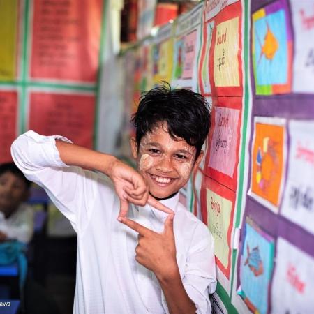 A boy uses his hands to create a square photo sign. He is in a classroom setting standing next to bulletin board with many colourful sheets pinned on it. He is wearing a white uniform shirt. In Cox's Bazaar, Bangladesh. 