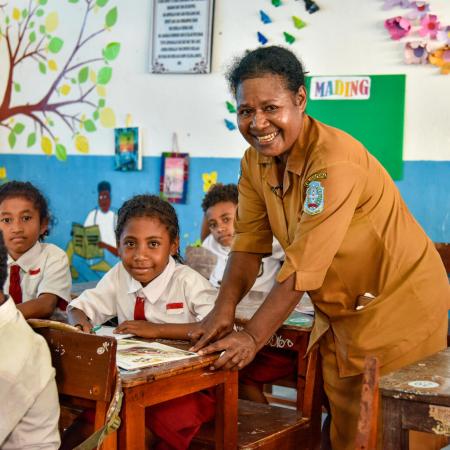 A teacher leans over a students desk and smiles at the camera. The student next to her is also smiling. Photo taken in Indonesia.