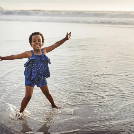 A young girl in stands in a body of water with her arms held wide smiling at the camera as the sun sets to the side of the photo