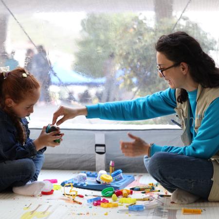 A child and a UNICEF staff member sit in a tent on the floor. The staff member giving the child a block with other toys lying around them.