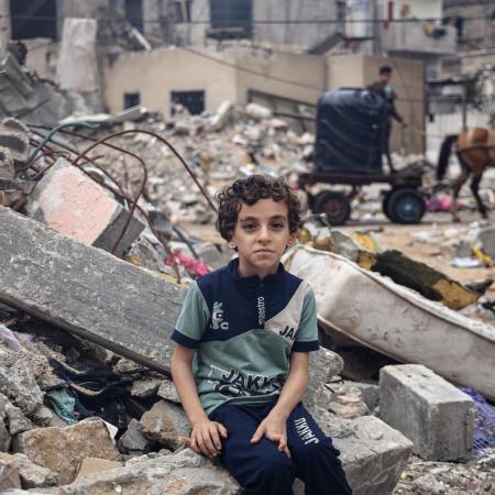 A child sits on the rubble of his family home, looking at the camera. There is all debris and rubble behind him. 