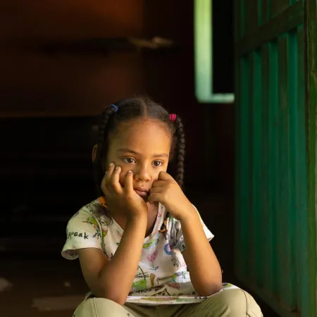 Young girl with pigtails, wearing a patterned shirt, sits pensively in a dimly lit doorway. She rests her chin on her hands, appearing thoughtful.