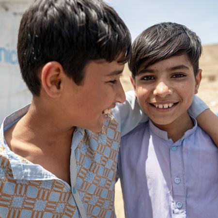 Two adolescent boys standing with their arms around each other’s shoulders and smiling outdoors in the sunshine, in front of a UNICEF tented shelter.