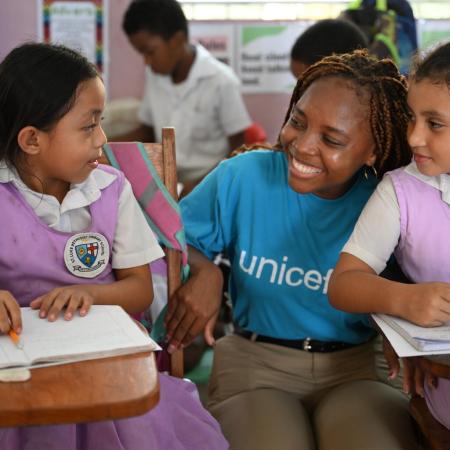 In a classroom, a woman wearing a blue UNICEF shirt crouch and smiles beside two girls sitting at their desk.