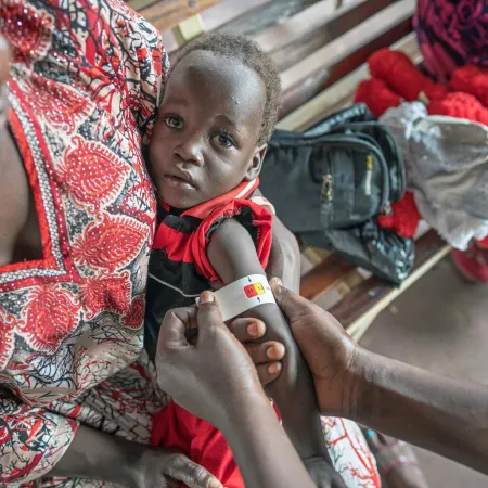 A caregiver holds a young child on her lap. The child wears a red and black outfit as a medical worker measures the child's upper arm with a MUAC tape to assess malnutrition.