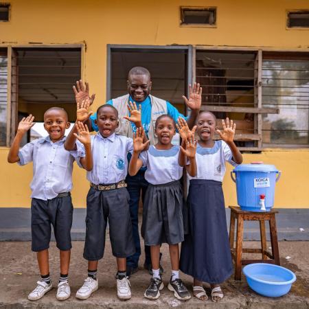 Un groupe d’enfants et un employé de l’UNICEF se tiennent à côté d’un seau d’eau, souriant à l’objectif tout en montrant leurs mains propres.