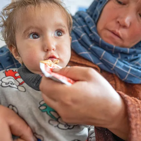 A baby eats a peanut paste out of a packet held by a caregiver in a blue headscarf.