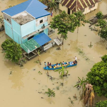 Individuals in a boat traversing a flooded street adjacent to a flooded house, highlighting the effects of severe flooding in the area.