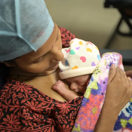 A mother wearing a surgical cap lovingly cradles her newborn, wrapped in a vibrant blanket. The baby wears a hat with colorful hearts, exuding warmth and care.