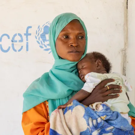  A woman in a green headscarf holds her baby in front of a wall with the UNICEF logo on it.