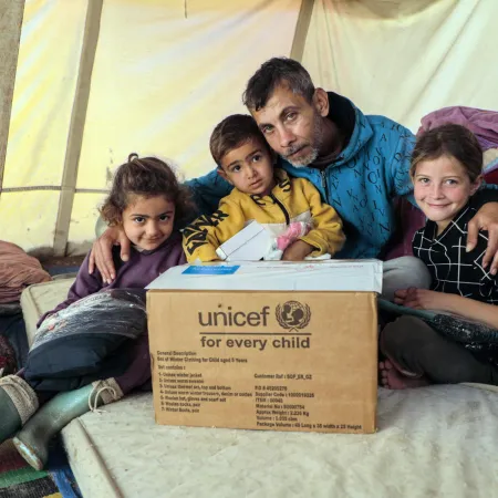 In an emergency tent, three children sit on the floor with their father who has his arms around them. In front of them is a UNICEF supply box.