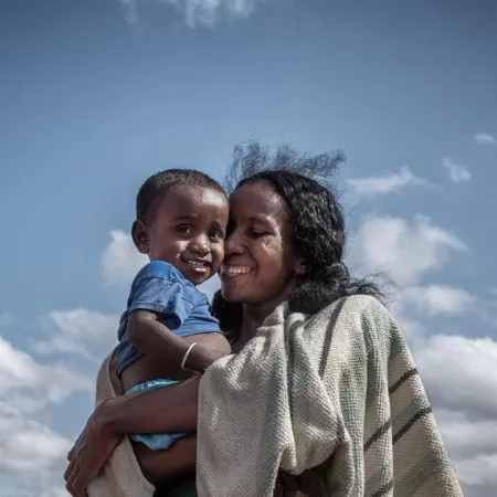 Une femme qui sourit tient dans ses bras un jeune enfant à l’extérieur, sous un ciel partiellement nuageux.