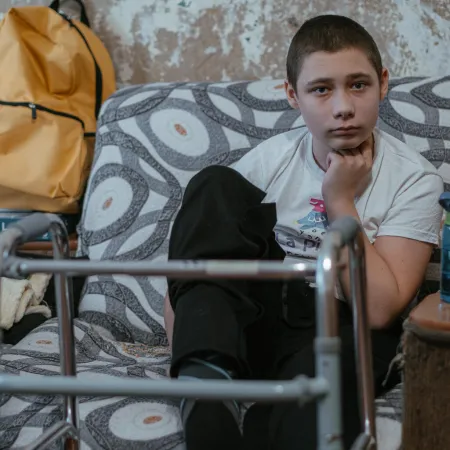 A boy sits on a patterned sofa, looking thoughtful. A walker is in front of him, a yellow backpack and hamster cage are nearby, and a bottle is on a table.