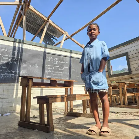 Smiling schoolgirl in a blue uniform stands in a classroom without a roof, surrounded by wooden desks and chalkboards under a clear sky.