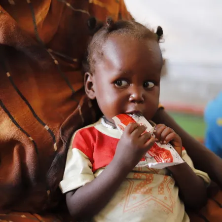 A small child sits on an adult's lap, holding a packet of nutritional paste to their mouth. The child's expression is thoughtful. The adult wears a patterned brown garment.