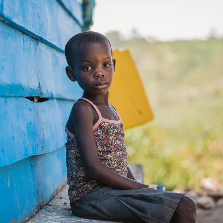A young child sits on a ledge beside a bright blue wooden wall, gazing thoughtfully at the camera. The background features a blurred, green landscape