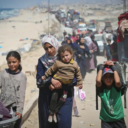 A woman carrying a child, a girl holding a basket and a boy wearing a backpack walk down a dusty road with other people.