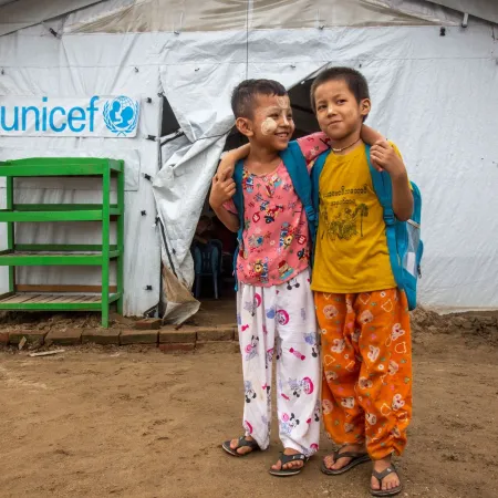 Deux enfants portant des sacs à dos se tiennent devant une tente temporaire de l’UNICEF, souriants, bras dessus bras dessous.