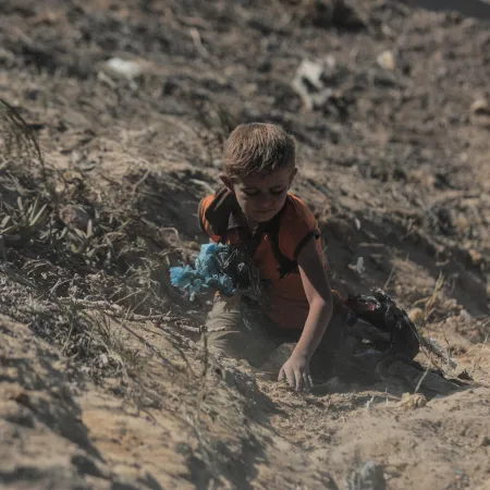 A young child crouches in a dry, dusty trench, reaching toward the ground while surrounded by scattered debris and sparse, scrubby plants. 