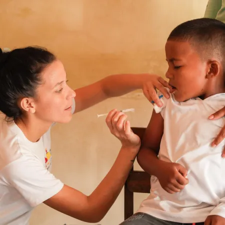 A healthcare worker administers a vaccine to a young boy sitting calmly, with an adult’s supportive hand on his shoulder. 
