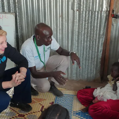 A woman in a UNICEF sweater and a man converse with a smiling woman holding a child, seated on mats in a metal-walled room, conveying warmth and hope.